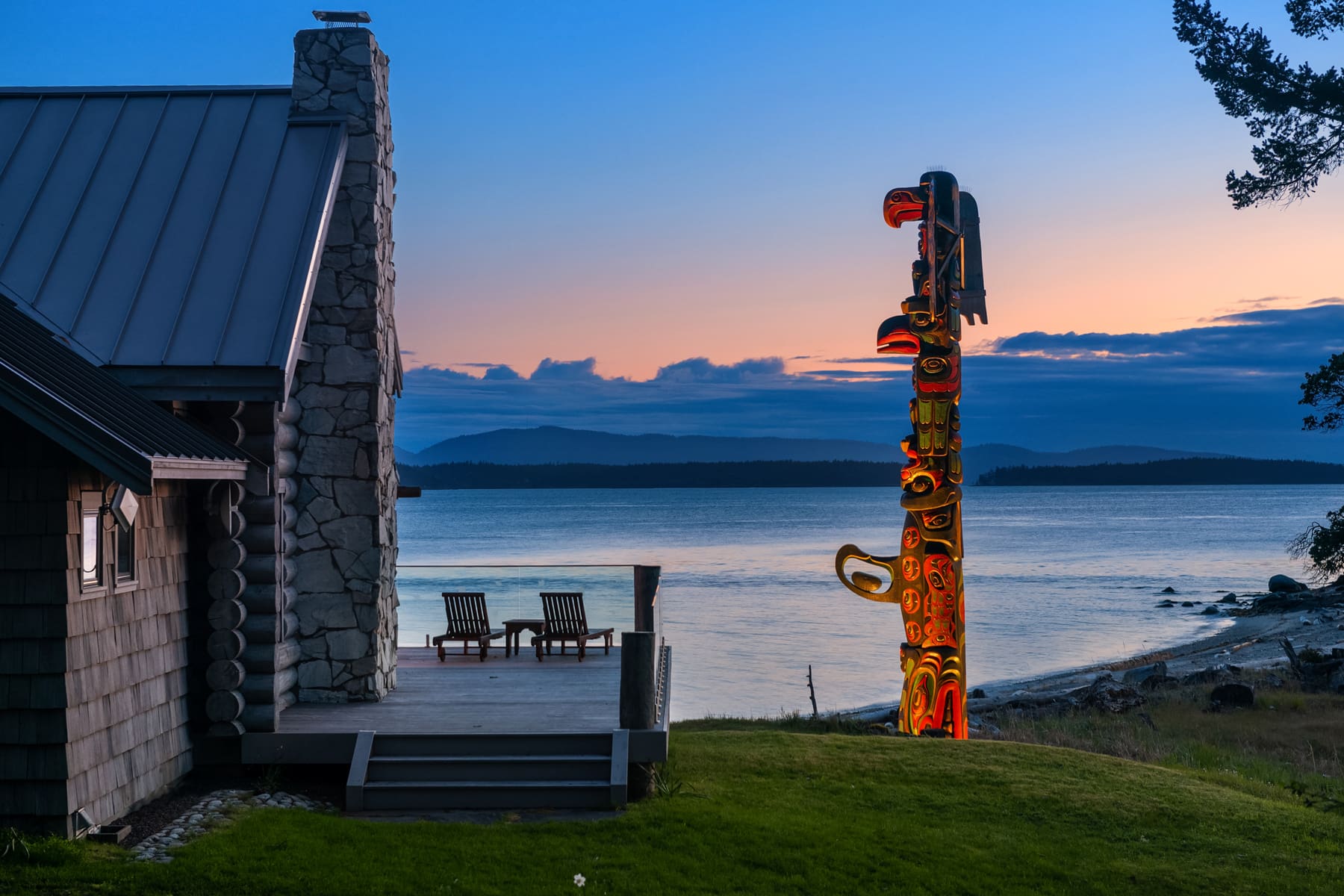 Cozy waterfront home exterior with a modern design, featuring a stone chimney and a stunning totem pole, set against a picturesque sunset over the Pacific Northwest.