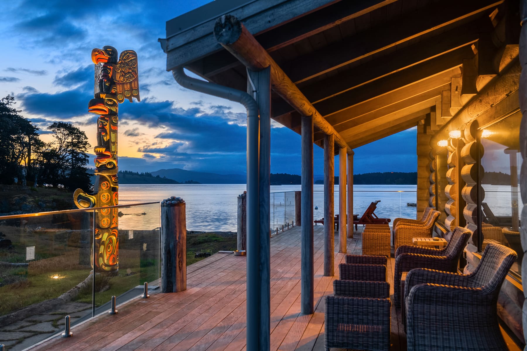 Cozy waterfront patio with wooden beams and wicker seating, showcasing a totem pole at dusk, located in the Pacific Northwest.