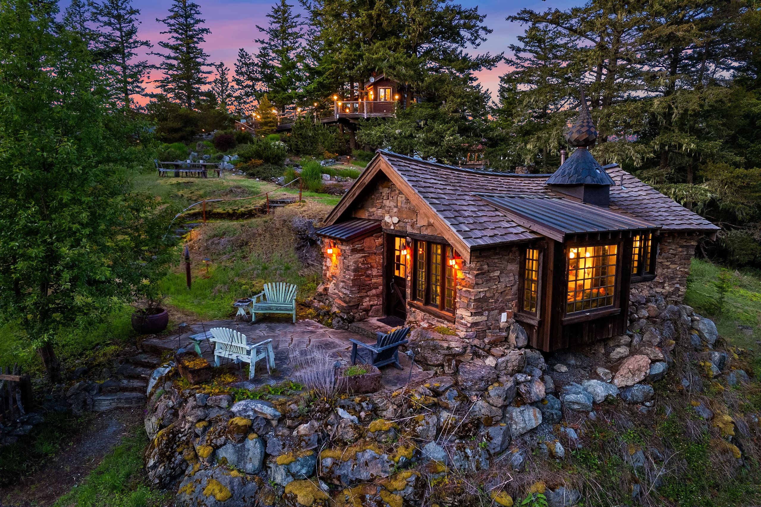 Charming stone cottage exterior with warm lighting, surrounded by lush greenery and rocky landscape, set against a twilight sky in the Pacific Northwest.