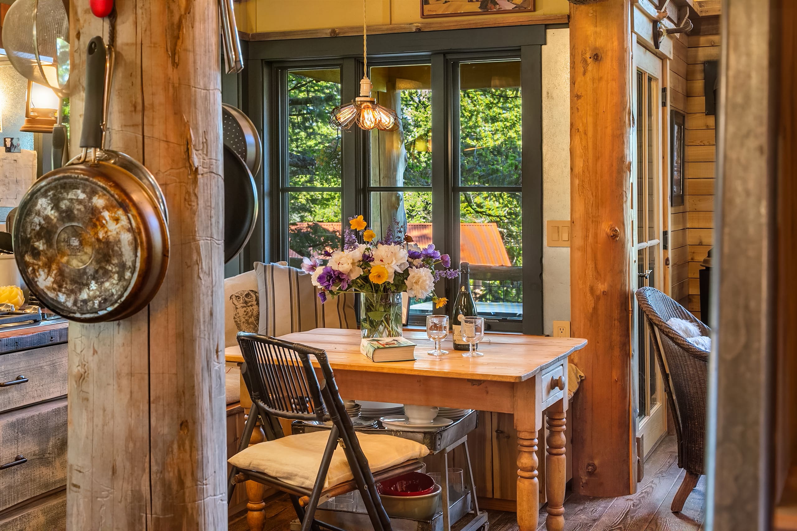 Cozy rustic kitchen dining area with a wooden table, vintage chairs, and a vibrant floral centerpiece, featuring warm lighting and large windows showcasing greenery, located in the Pacific Northwest.