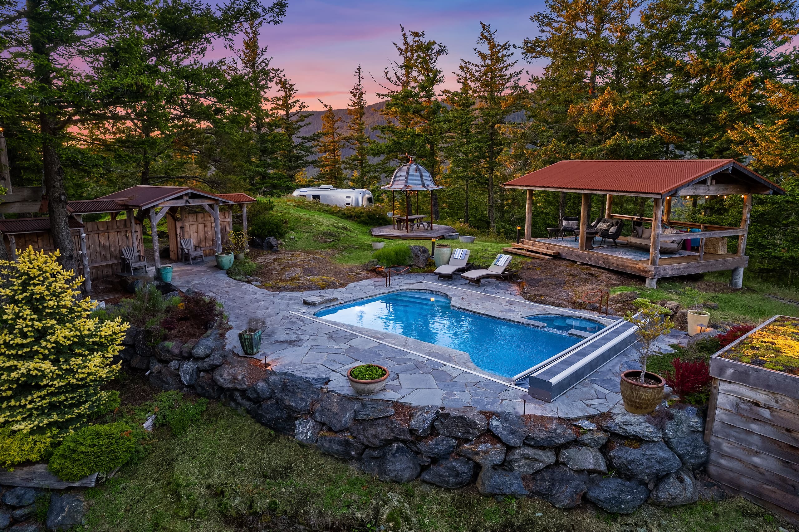 Outdoor oasis featuring a modern pool surrounded by natural stone landscaping, cozy seating areas, and a rustic gazebo, set against a serene forest backdrop in the Pacific Northwest.