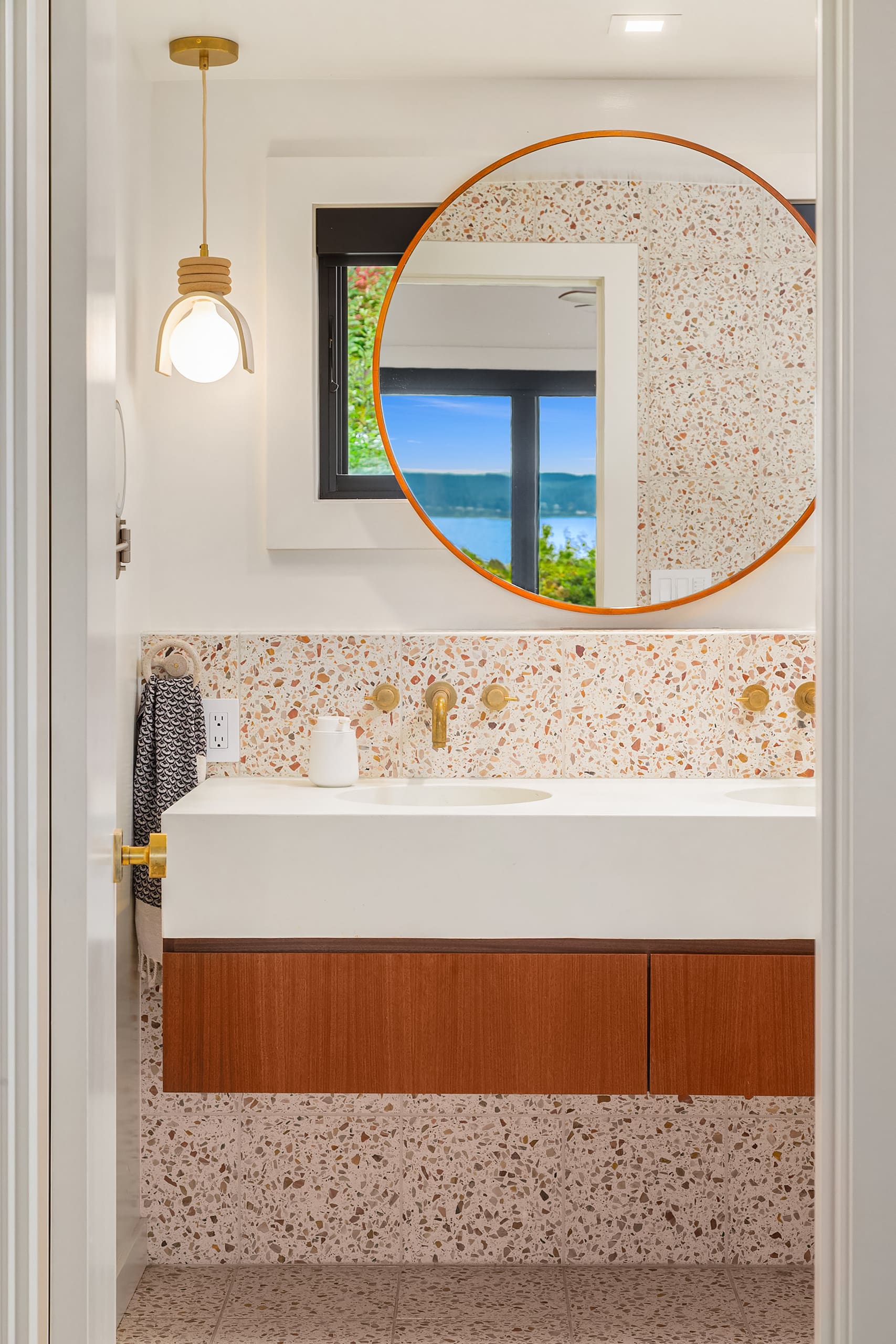 Modern bathroom featuring a sleek double vanity with warm wood accents, terrazzo wall tiles, and a large circular mirror. Bright and inviting atmosphere with a view of the Pacific Northwest landscape visible through the window.