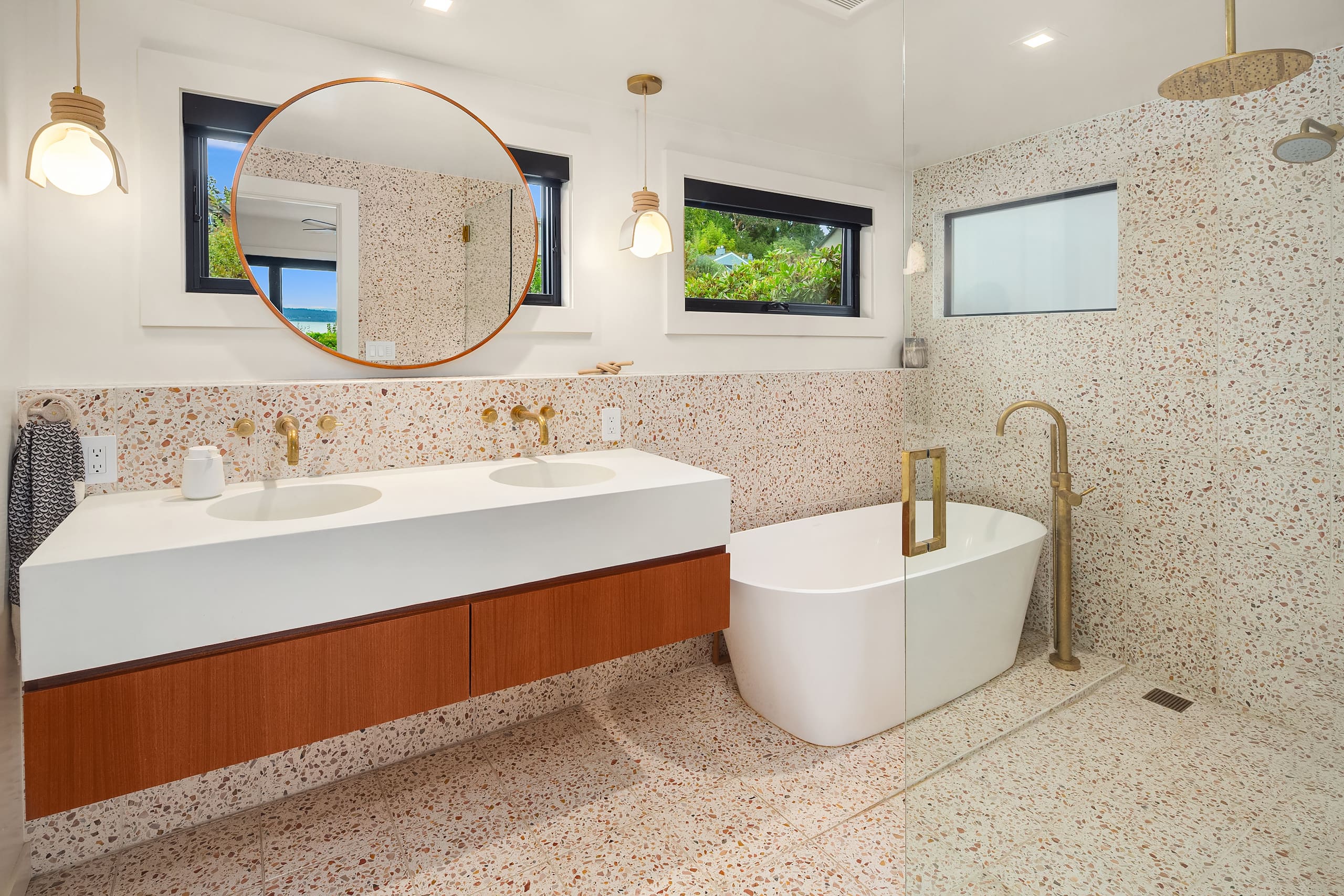 Modern bathroom featuring a double sink vanity with a sleek white countertop, wooden cabinetry, and a freestanding tub. The space is adorned with terrazzo walls, minimalist lighting, and large windows providing natural light. Located in the Pacific Northwest.