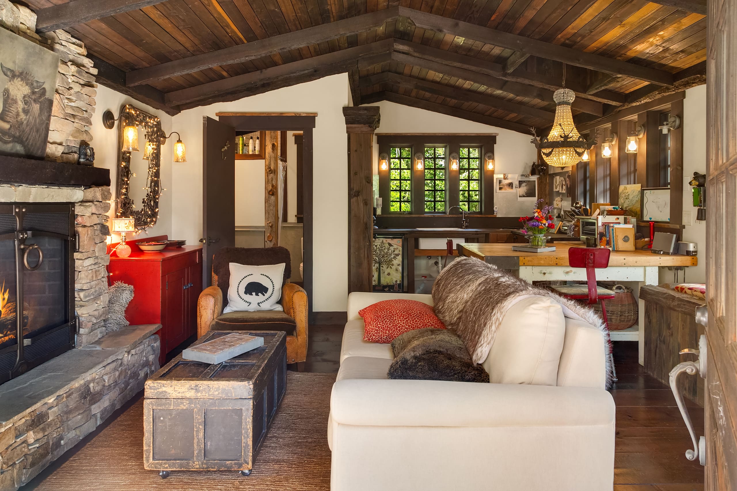Cozy living room in a rustic cabin featuring exposed wooden beams, a stone fireplace, and warm lighting. The space includes a comfortable sofa, vintage coffee table, and a charming kitchen area, located in the Pacific Northwest.