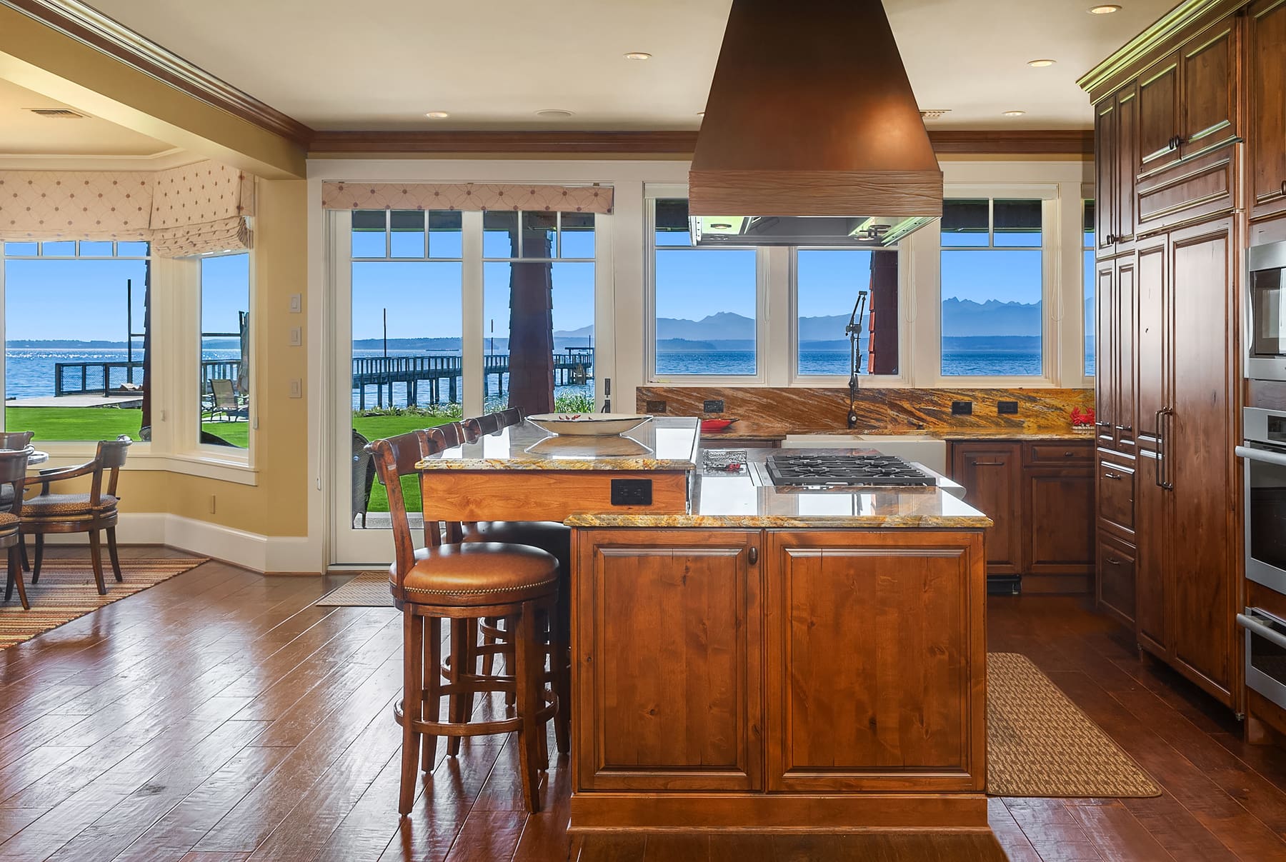 Spacious modern kitchen with stunning water views, featuring rich wooden cabinetry, a large island with a gas cooktop, and elegant finishes, located in the Pacific Northwest.