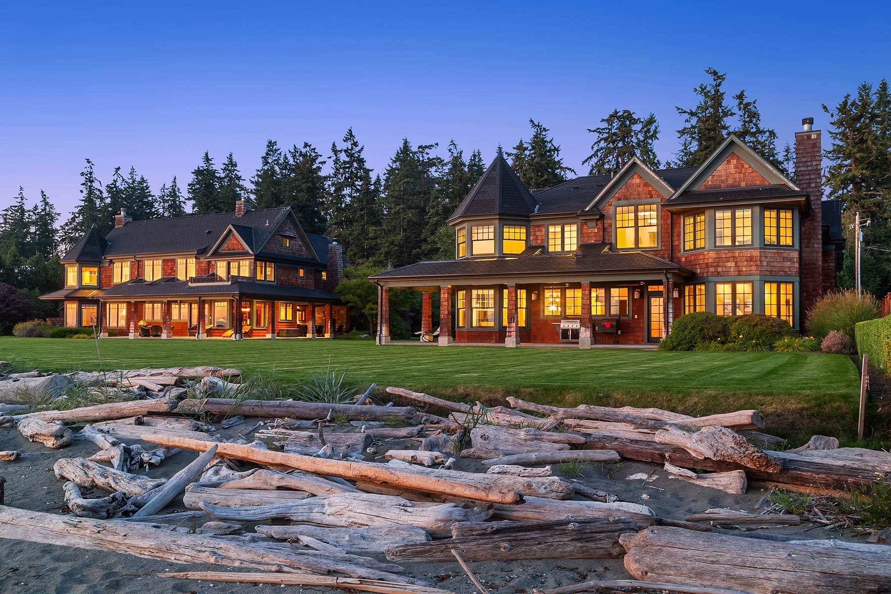 Stunning exterior view of a luxurious waterfront property at twilight in the Pacific Northwest, showcasing elegant architecture with large windows and a manicured lawn, surrounded by lush trees and driftwood along the beach.