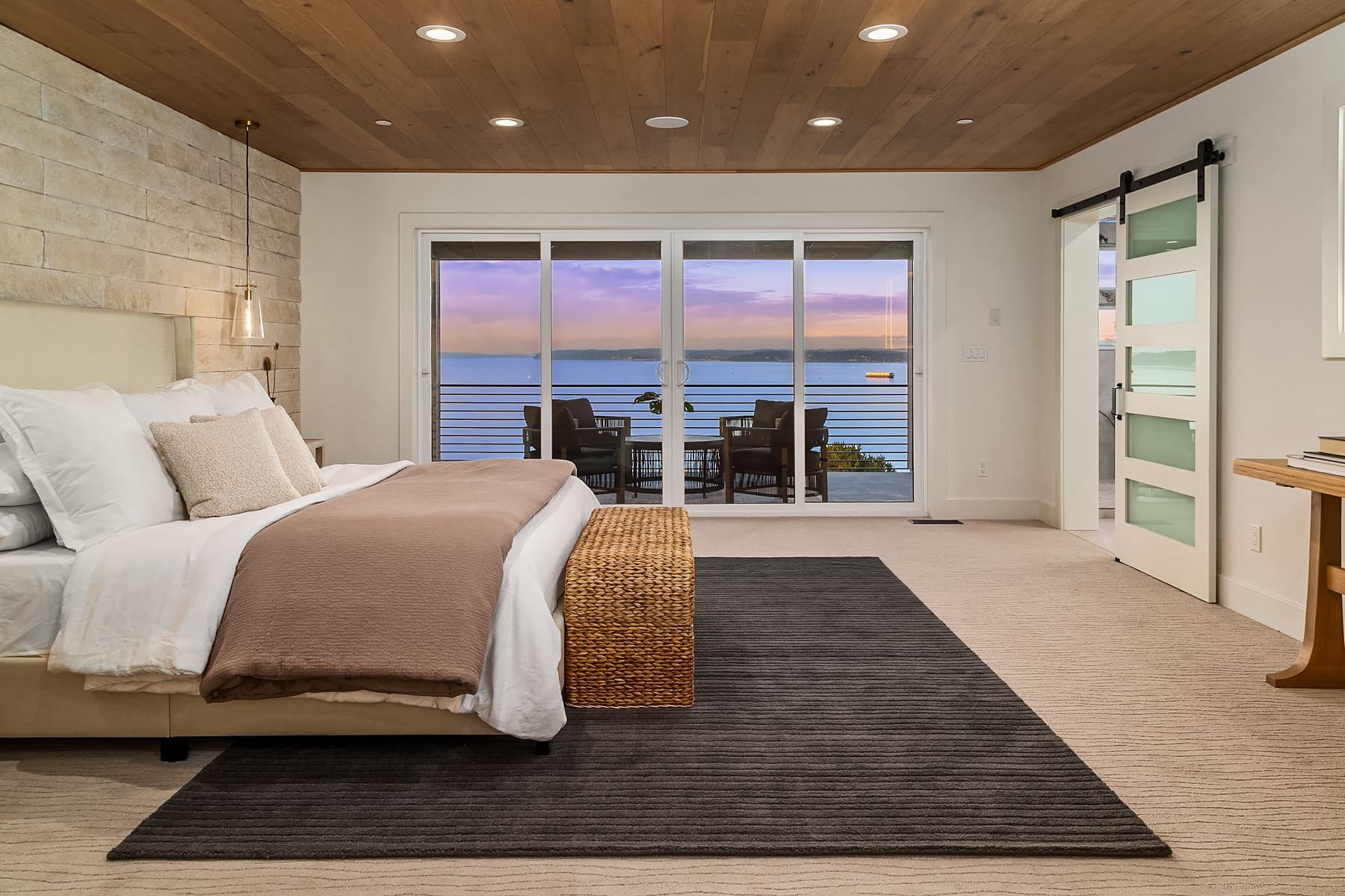 Modern bedroom with a serene waterfront view, featuring a cozy bed with neutral bedding, warm wood accents, and stylish sliding glass doors opening to a private balcony in the Pacific Northwest.