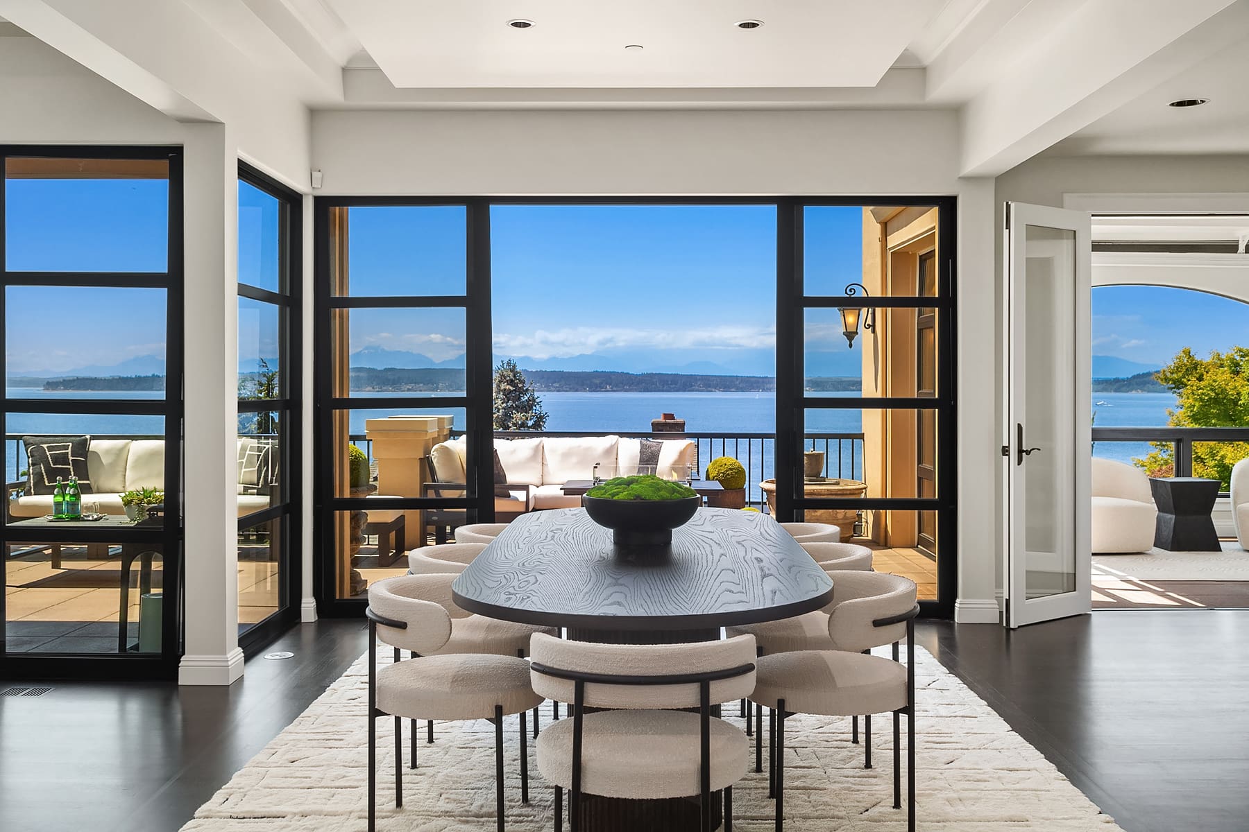 Modern dining area with an elegant design featuring a large black table surrounded by plush cream chairs, overlooking a stunning waterfront view in the Pacific Northwest. Bright, airy space with floor-to-ceiling windows and seamless indoor-outdoor flow.