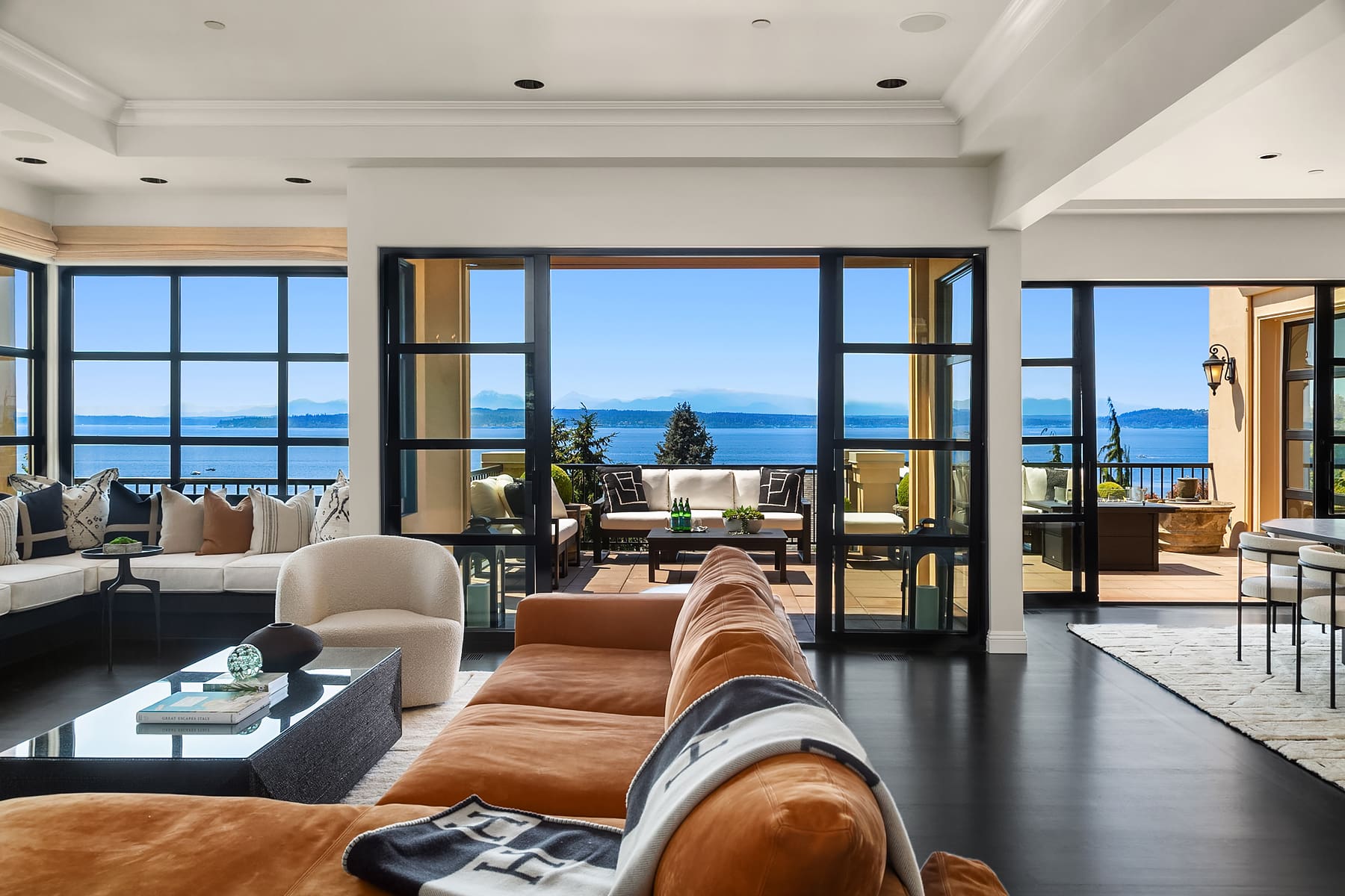 Spacious modern living room with panoramic views of the water and mountains, featuring a cozy sectional sofa, stylish decor, and large glass doors opening to an elegant outdoor seating area, located in the Pacific Northwest.