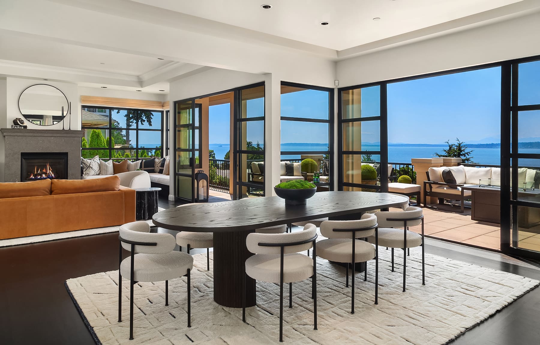 Modern open-concept living and dining area featuring a stylish round table with upholstered chairs, a cozy sofa, and large glass doors offering stunning views of the water, located in the Pacific Northwest.