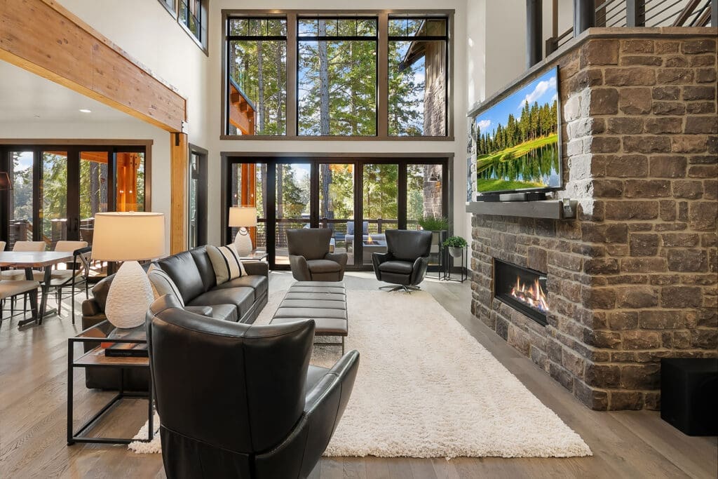 Modern living room with large windows showcasing a forest view, featuring a cozy fireplace, stylish black leather furniture, and warm wood accents, located in the Pacific Northwest.