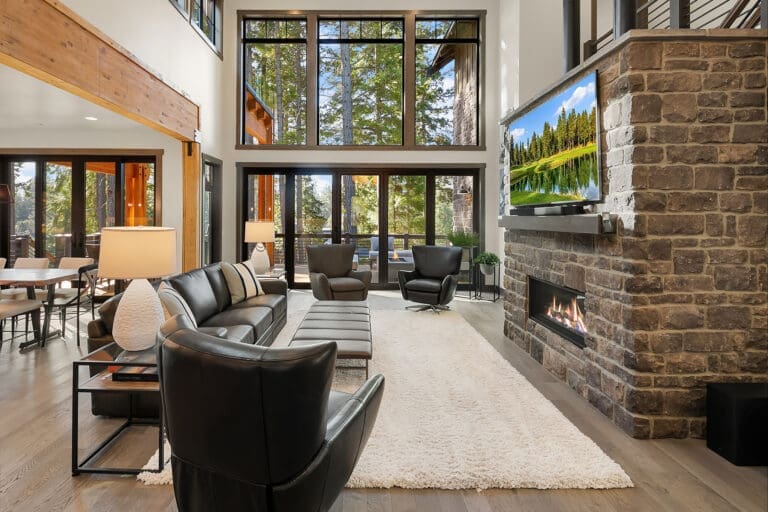 Modern living room with large windows showcasing a forest view, featuring a cozy fireplace, stylish black leather furniture, and warm wood accents, located in the Pacific Northwest.