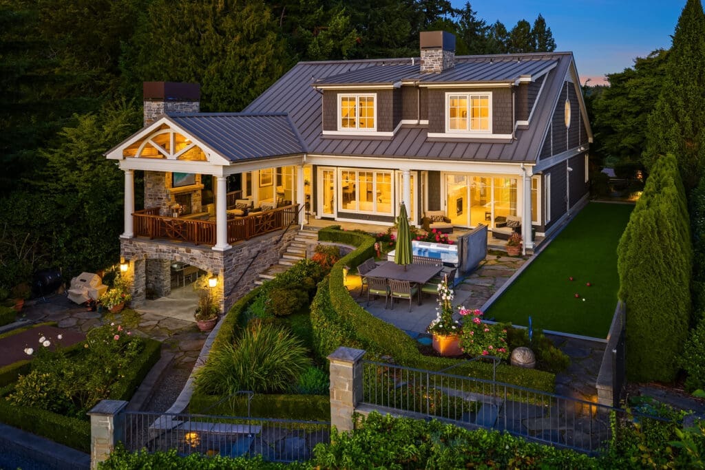 Medina's first mansion. An elegant exterior of a modern home in the Pacific Northwest, featuring stone accents, a spacious patio with outdoor seating, and beautifully landscaped gardens, illuminated at twilight.