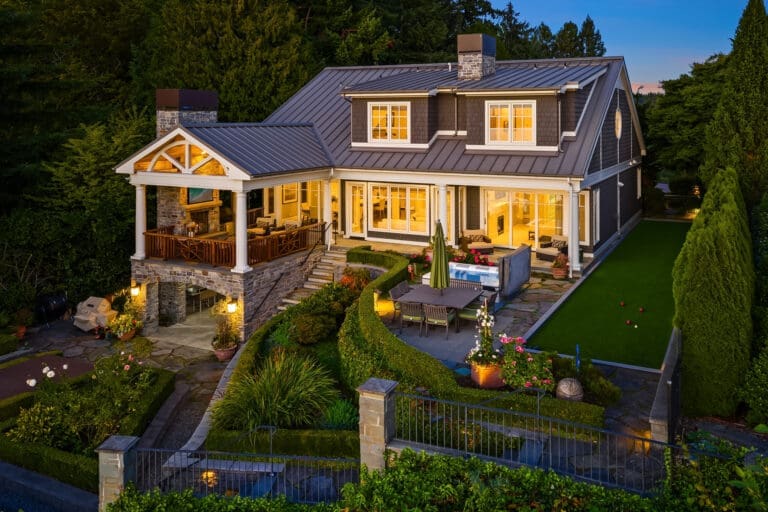 Medina's first mansion. An elegant exterior of a modern home in the Pacific Northwest, featuring stone accents, a spacious patio with outdoor seating, and beautifully landscaped gardens, illuminated at twilight.