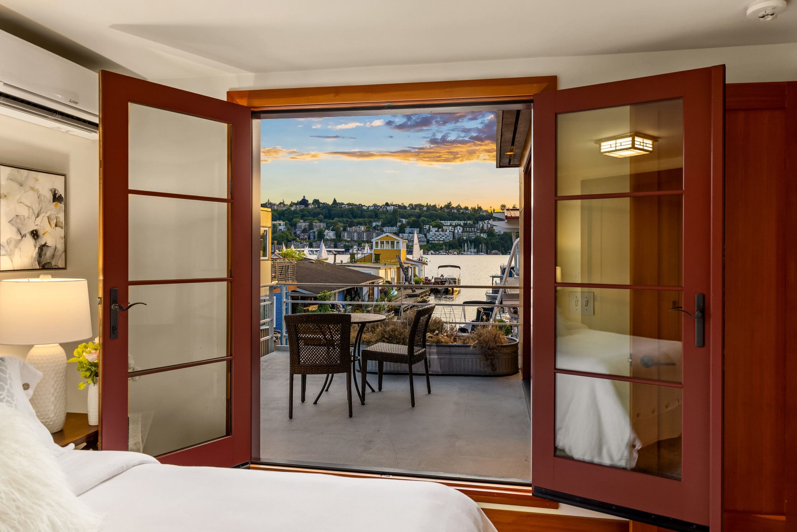 Cozy bedroom with open double doors leading to a scenic balcony overlooking Lake Union at sunset, featuring modern furnishings and a tranquil atmosphere, located in Seattle.