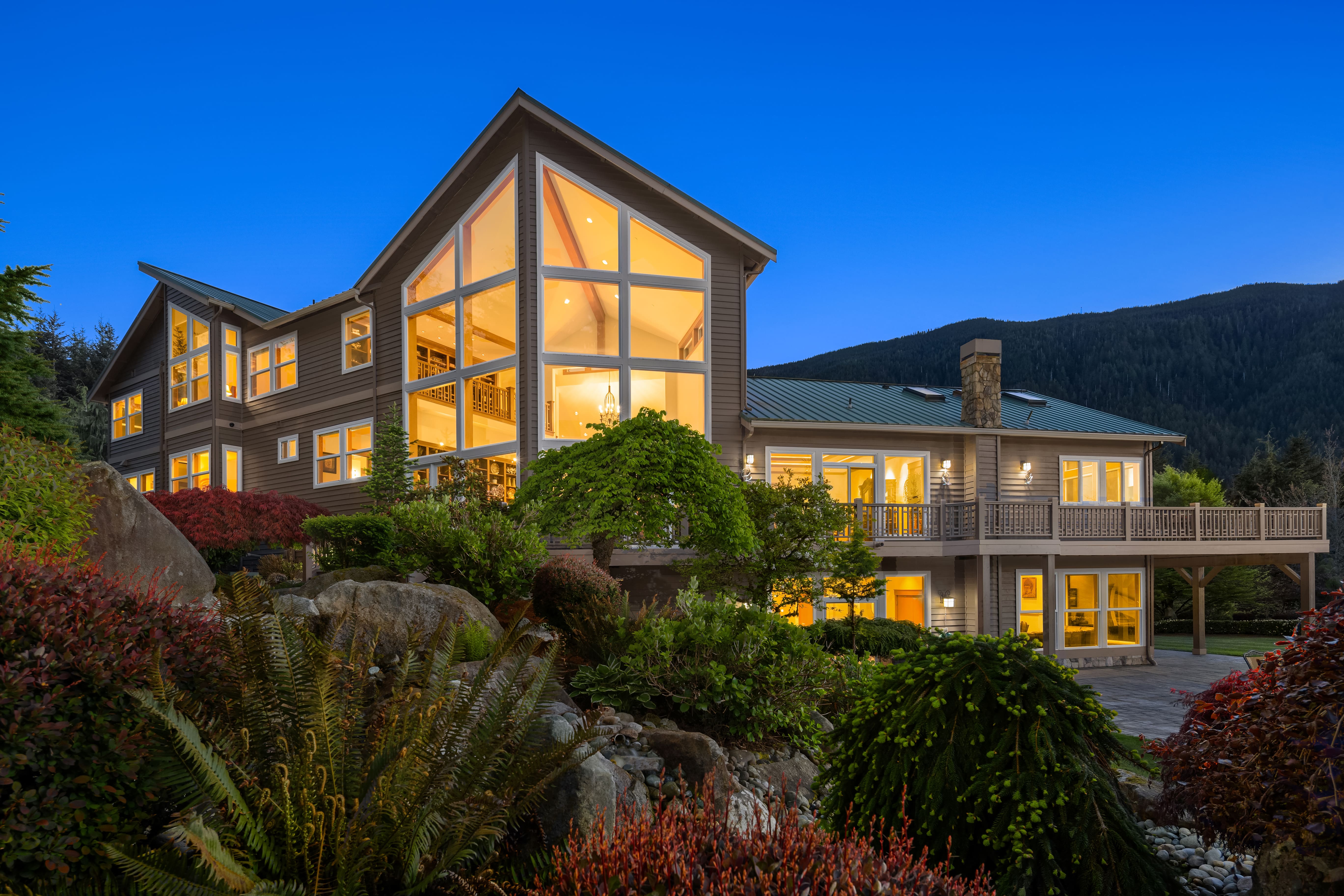 Elegant exterior view of a modern home surrounded by lush landscaping in the Pacific Northwest at twilight, showcasing large windows that illuminate the design and mountain backdrop.